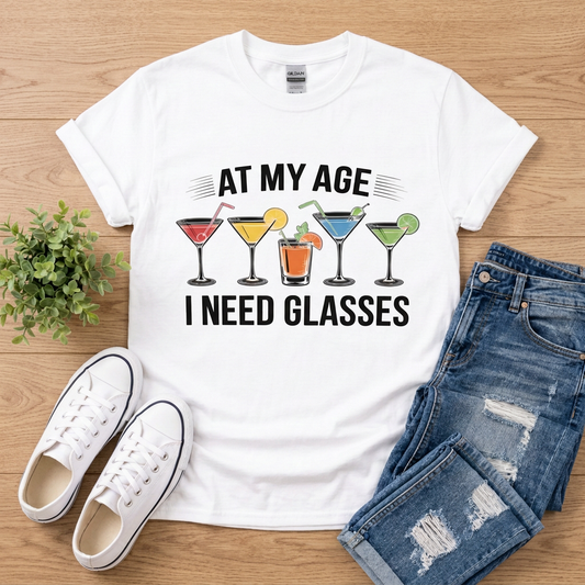 White At My Age I Need Glasses cocktail graphic T-shirt lying flat on brown wood with jeans and white sneakers
